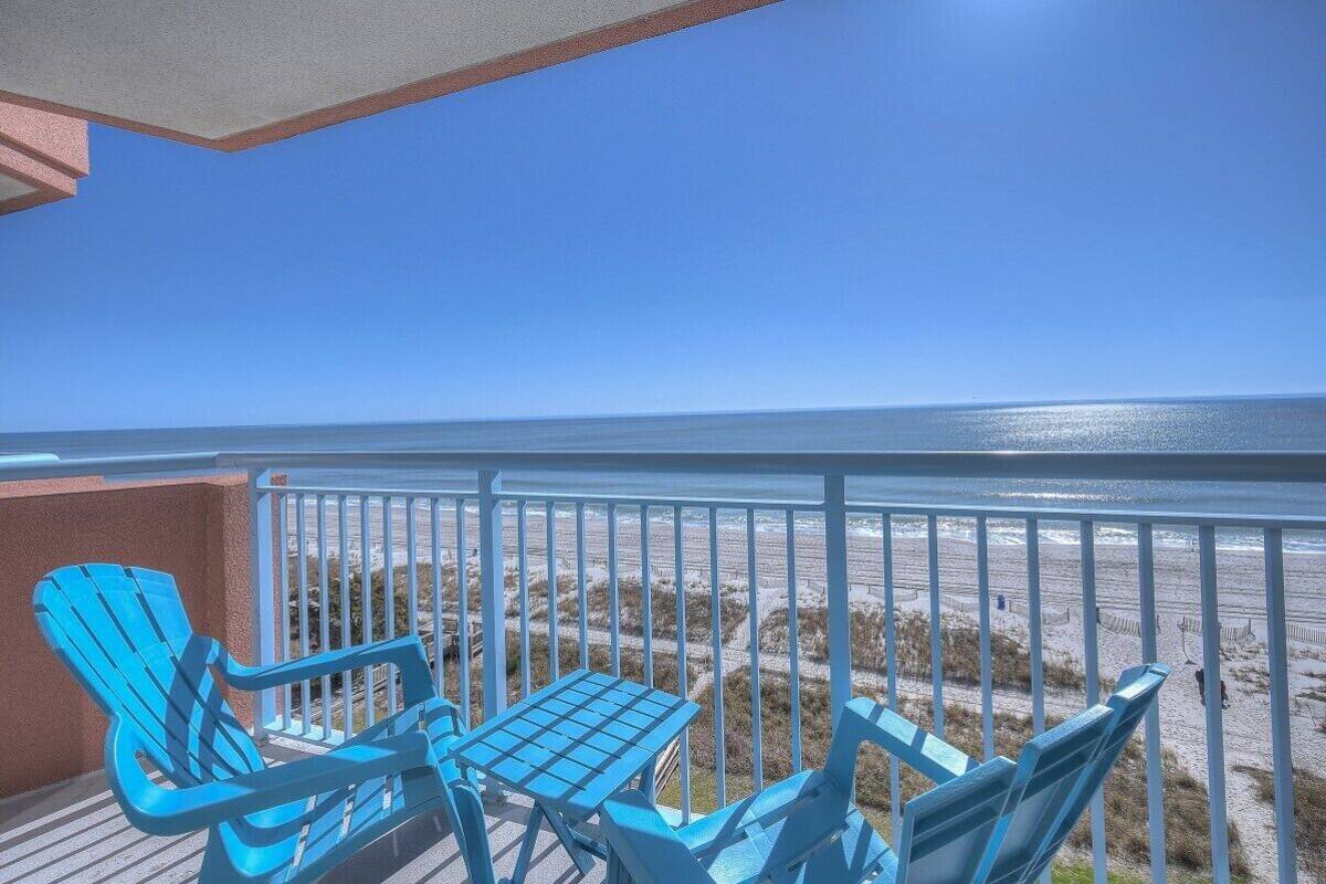 Ein Balkon mit blauen Adirondack-Stühlen und einem kleinen Tisch, mit Blick auf einen Sandstrand und den Ozean.