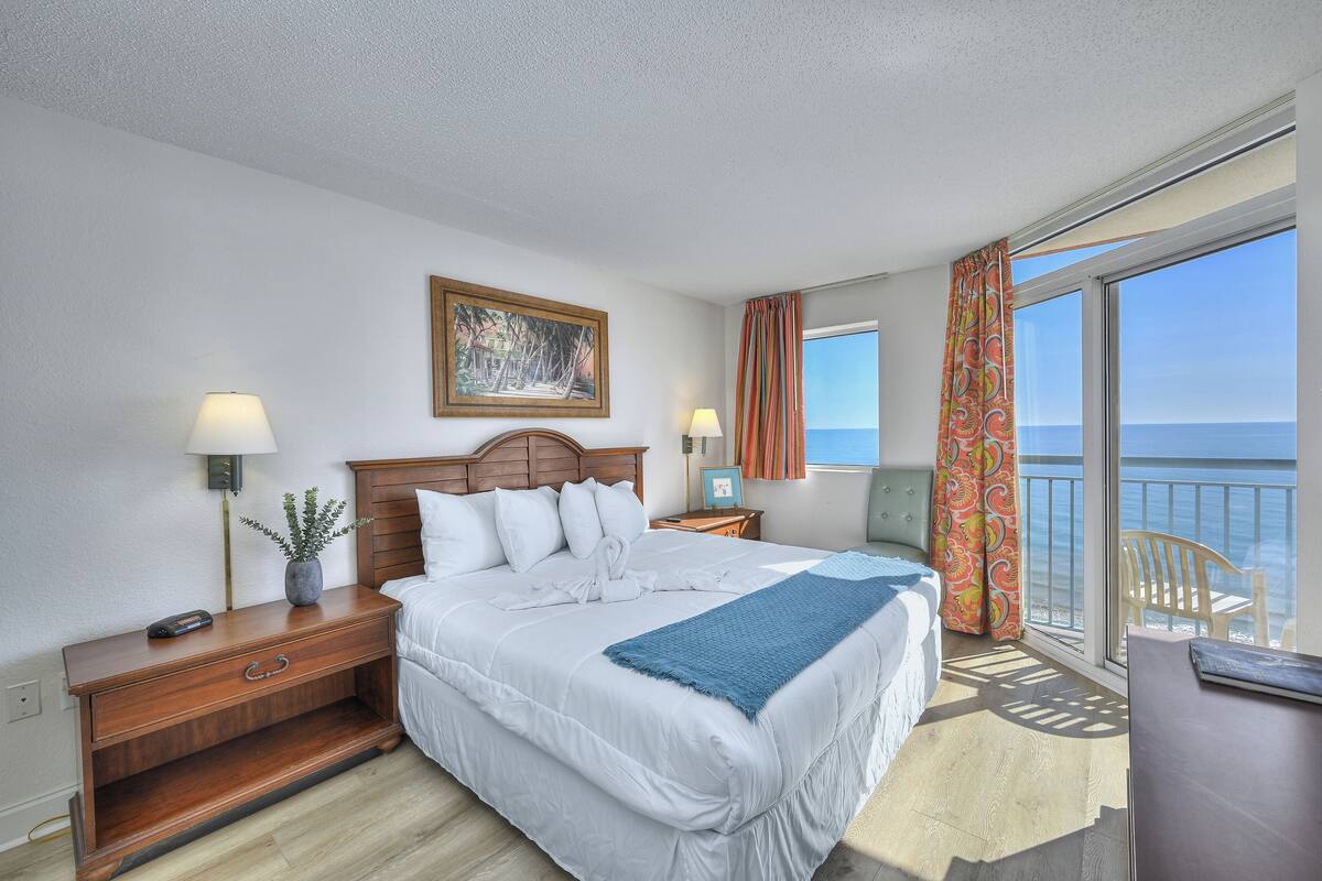 Ein Hotelzimmer mit einem großen Bett, einem Nachttisch aus Holz, einem Schreibtisch und Meerblick vom Balkon.