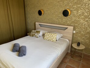 Een net opgemaakt bed met kussens, een rond bijzettafeltje met een vaas, en een muur met twee ronde wandlampen.