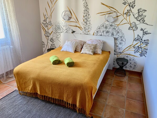 Een net opgemaakt bed met een gele deken en twee groene handdoeken, een wit hoofdeinde en een muur met bloemen- en bladmotieven.