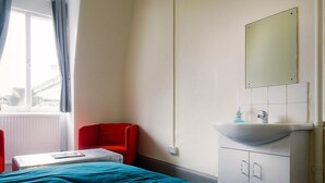 Una habitación de hotel con cama, una silla roja, una ventana con cortinas, un lavamanos blanco y un espejo.