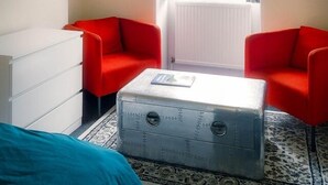 Una habitación con un ventanal grande, dos sillas rojas, un tocador blanco y una maleta antigua.