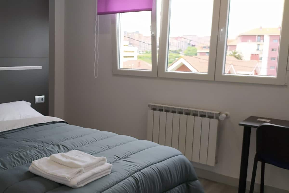 A bedroom with a bed, a window with a view of buildings, a radiator, and a small table.