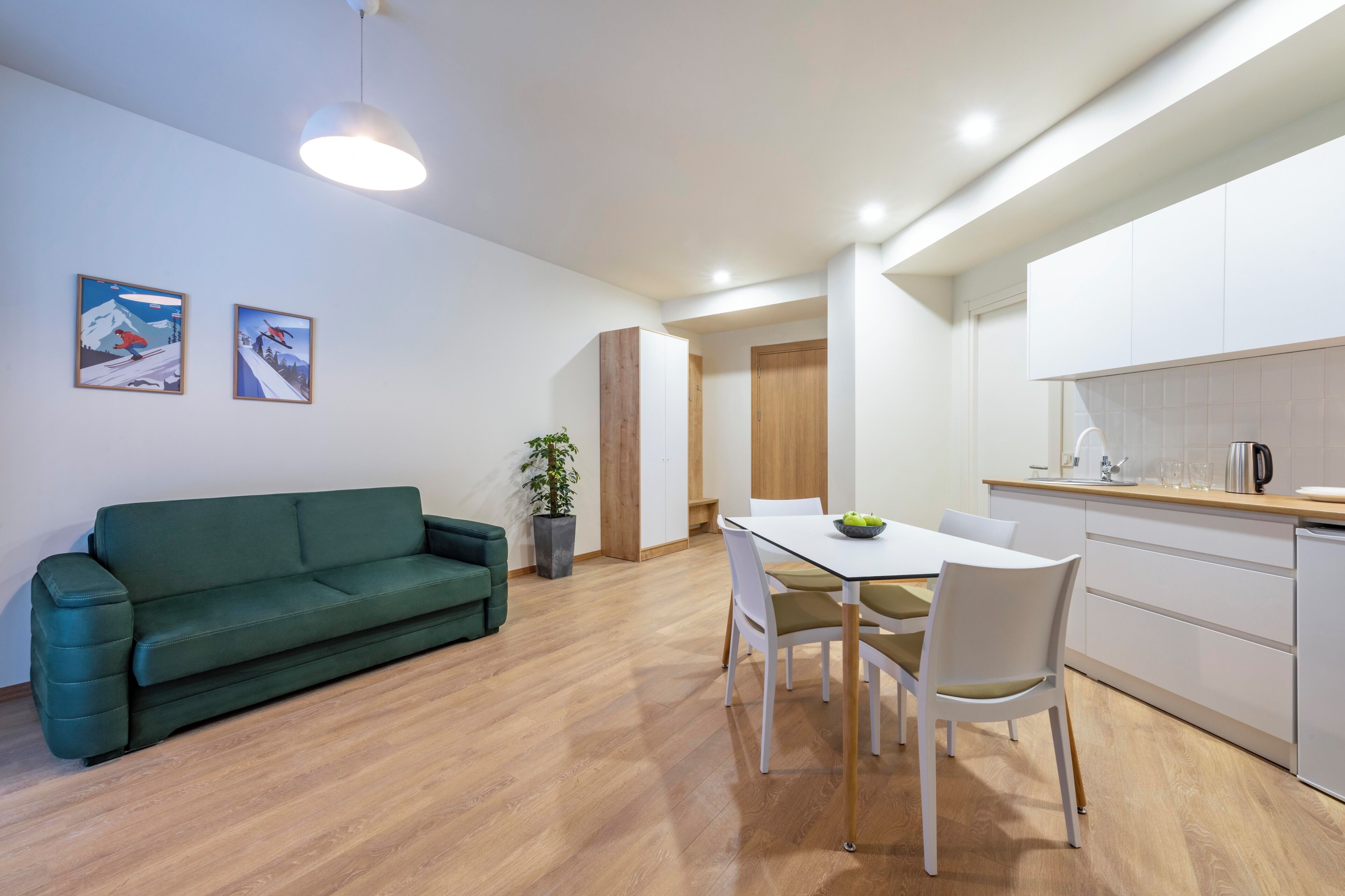 A modern living room with a green sofa, a dining table with chairs, a potted plant, and a kitchen area with white cabinets and a countertop.