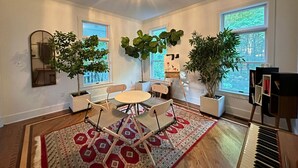 Une pièce spacieuse avec une grande fenêtre, un piano, une table ronde entourée de chaises et deux plantes en pot.