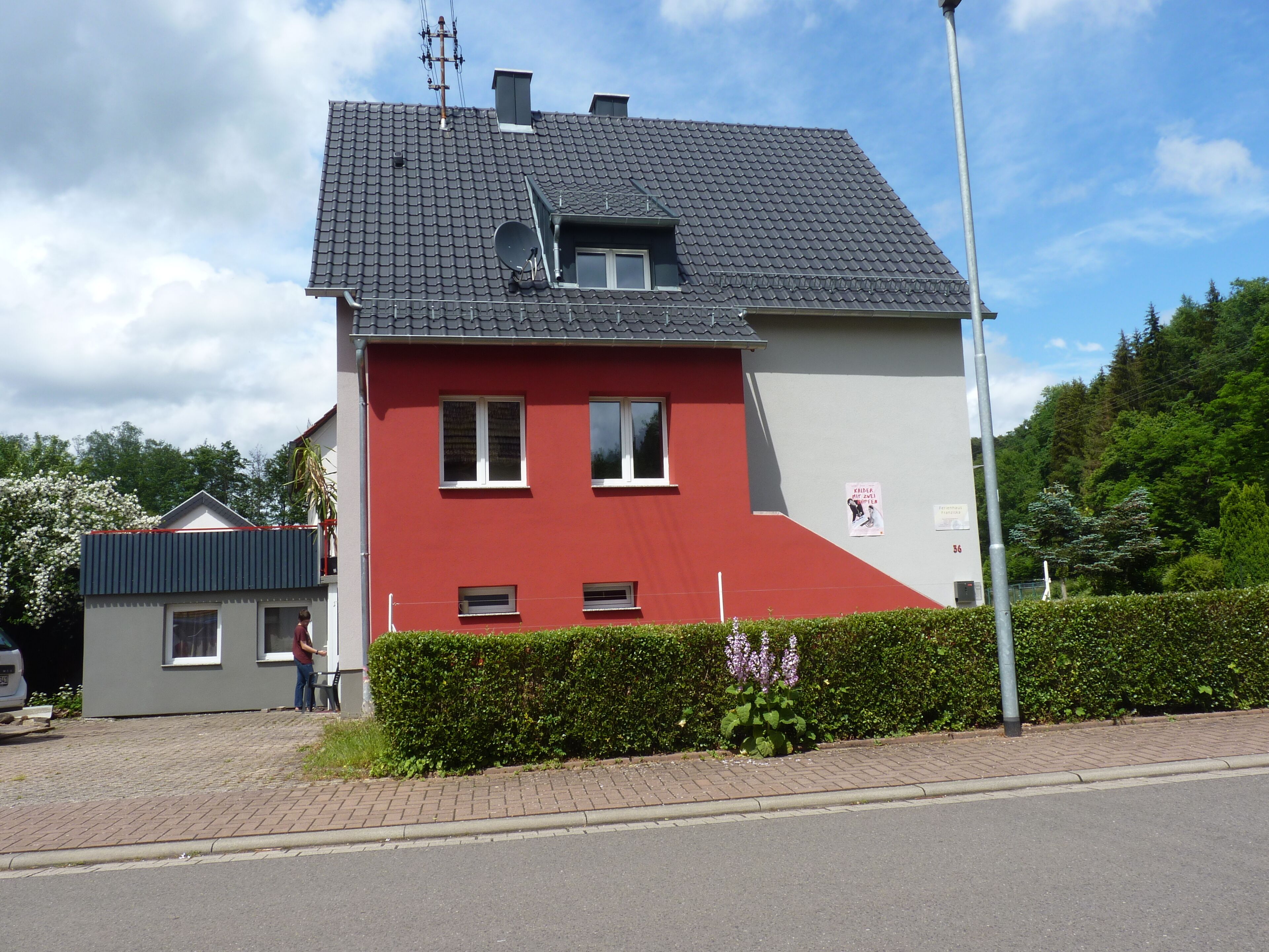 A two-story house with a red and grey facade, a satellite dish, and a small garden with flowers.