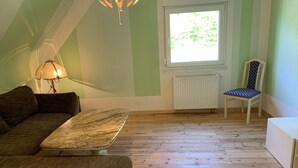Een kamer met een houten vloer, een bank, een tafel, een stoel, een lamp en een raam met uitzicht op groen.