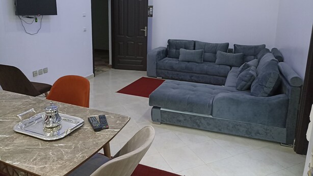 Una sala de estar con un sofá gris, un televisor montado en la pared y una mesa de comedor con una bandeja y controles remotos.