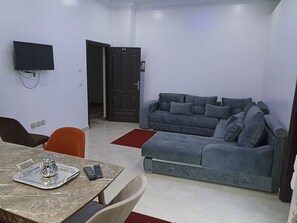 Een woonkamer met een grijze bank, een aan de muur bevestigde televisie en een eettafel met een dienblad en afstandsbedieningen.