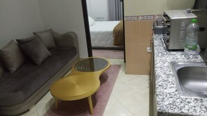 Un espacio habitable compacto con sofá, una mesita redonda, una kitchenette con pileta y una botella de agua, y una cama al fondo.