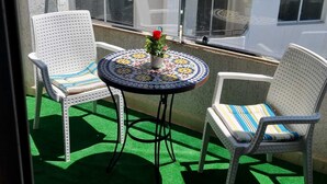 Un balcón con dos sillas blancas de mimbre, una mesita redonda con una cubierta de mosaico de colores y una maceta con una flor roja.