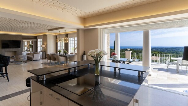 Una cucina moderna con un'isola grande, un'area pranzo con un tavolo in vetro e un balcone con vista sul paesaggio.