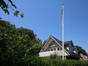 A house with a prominent flagpole, surrounded by dense greenery and under a clear blue sky.