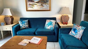 Una sala de estar con un sofá azul, dos lámparas, un cuadro y una mesa de centro con una revista.
