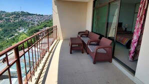 Un balcón con sofá y mesa, vista a un paisaje urbano y una ventana con cortinas.