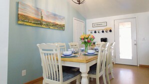 A dining area with a table set for four, surrounded by white chairs with blue cushions. A chandelier hangs from the ceiling, and a painting of a landscape is on the wall. - Cozy Swell House- Pet Friendly, Fenced Backyard: 2 King Beds (Page)