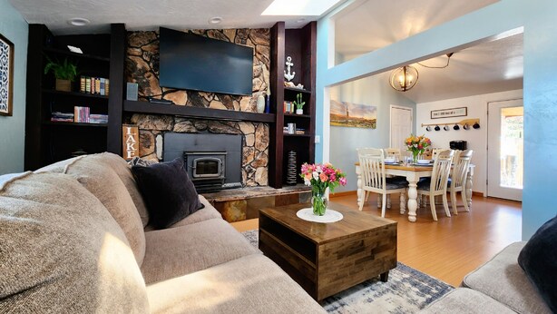 A living room with a stone fireplace, a flat-screen TV, a dining area with a table and chairs, and a bookshelf. - Cozy Swell House- Pet Friendly, Fenced Backyard: 2 King Beds (Page)