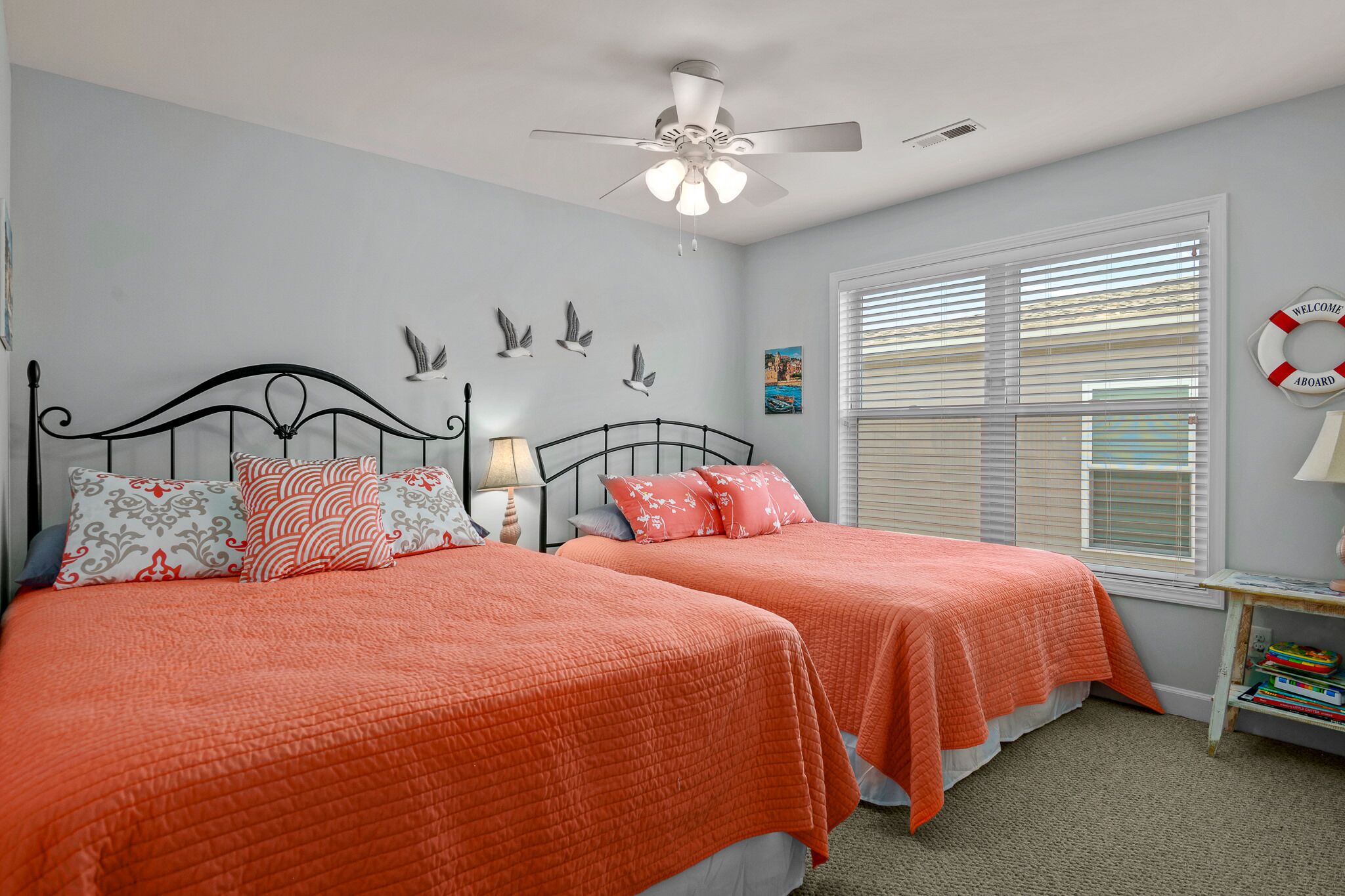 A bedroom with two beds, a ceiling fan, a window with blinds, and a small table with books.