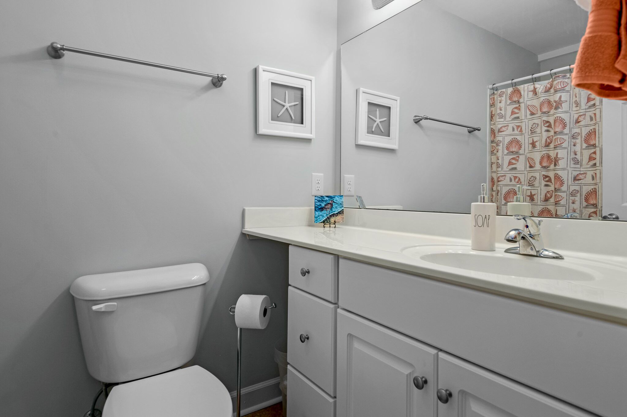 A bathroom with a toilet, a sink, and a mirror. There are two framed pictures on the wall, a towel rack, and a shower curtain with a shell pattern.
