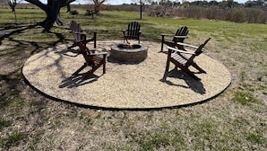 Uma área de estar externa circular com cadeiras Adirondack e uma lareira, cercada por cascalho e grama, com árvores ao fundo.