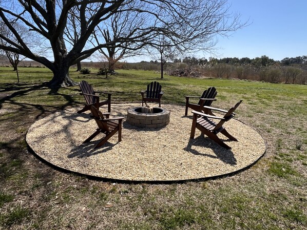 A circular outdoor seating area with Adirondack chairs and a fire pit, surrounded by gravel and grass, with trees in the background.