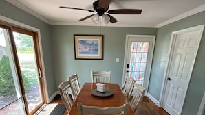 Uma sala de jantar com mesa de madeira, cadeiras brancas, ventilador de teto e um quadro na parede.