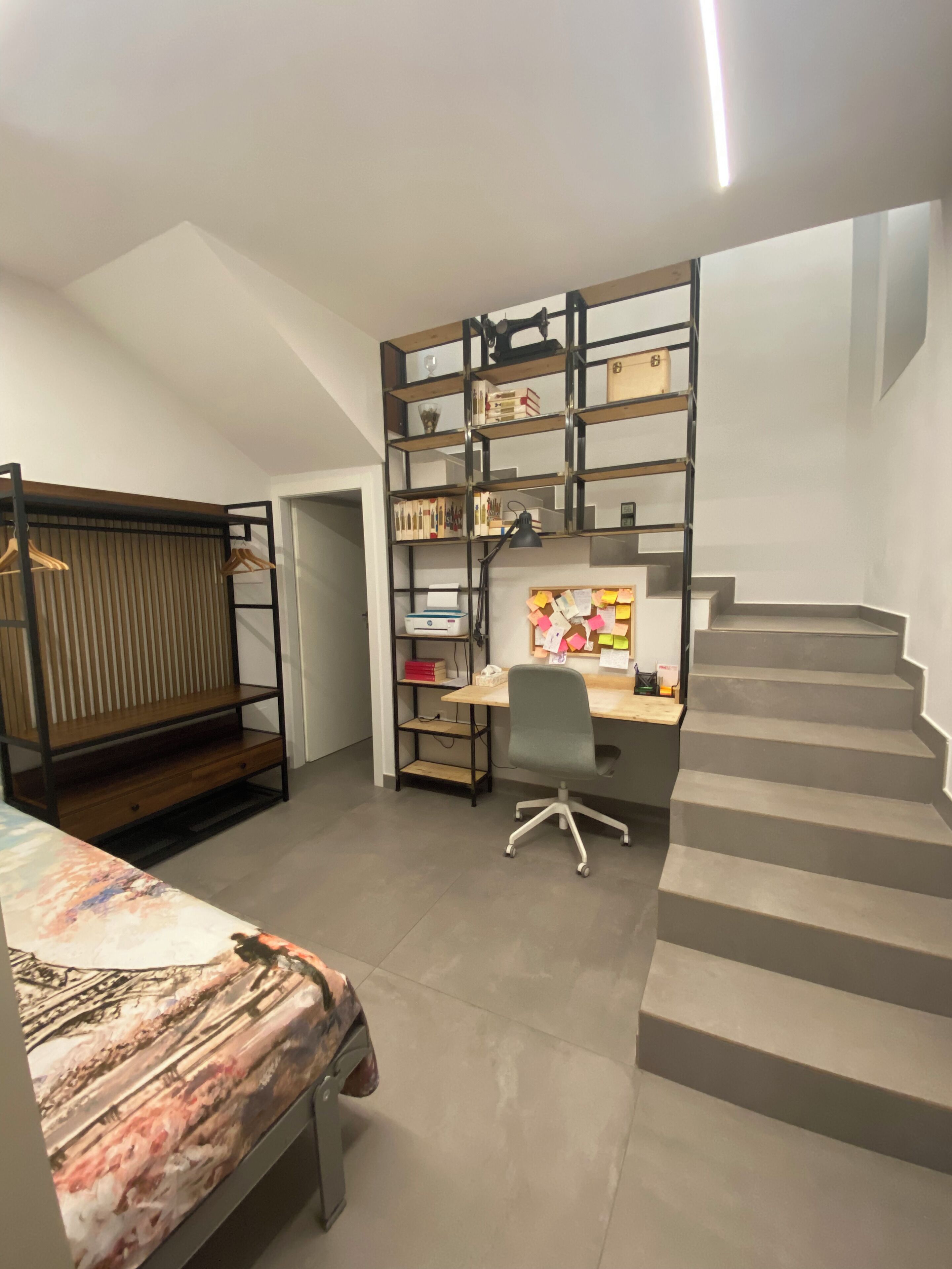 A bedroom with a bed, a desk with a chair, a bookshelf, and a staircase.