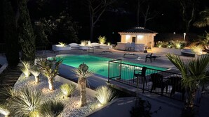 Une piscine avec une terrasse carrelée, entourée de palmiers et de plantes, et un kiosque en arrière-plan.