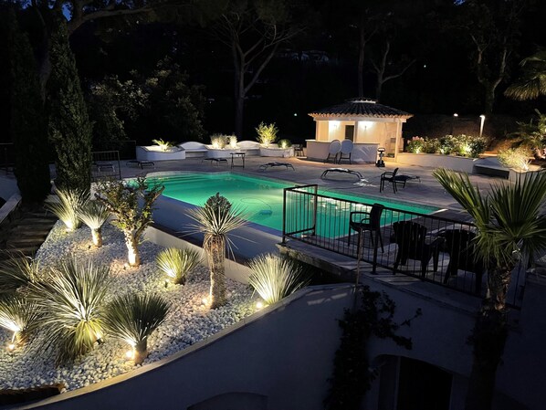 A swimming pool with a tiled deck, surrounded by palm trees and plants, and a gazebo in the background.