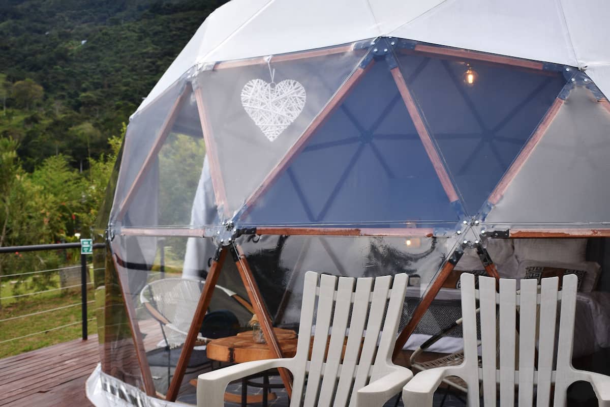 A geodesic dome structure with a transparent roof, two white chairs, and a wooden deck.