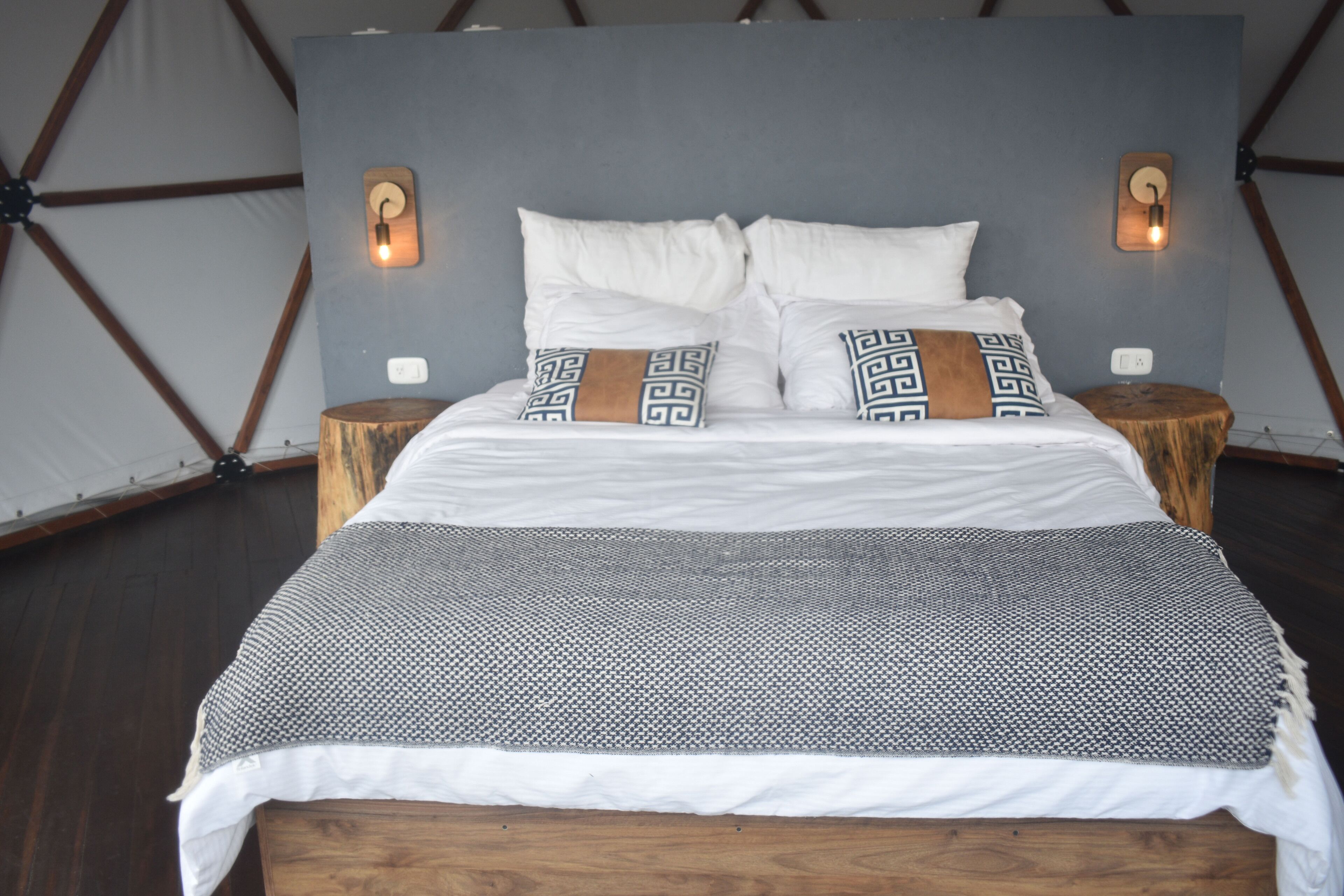 Una cama con almohadas blancas y estampadas, una manta gris y negra, y cabecera y mesitas de noche de madera.