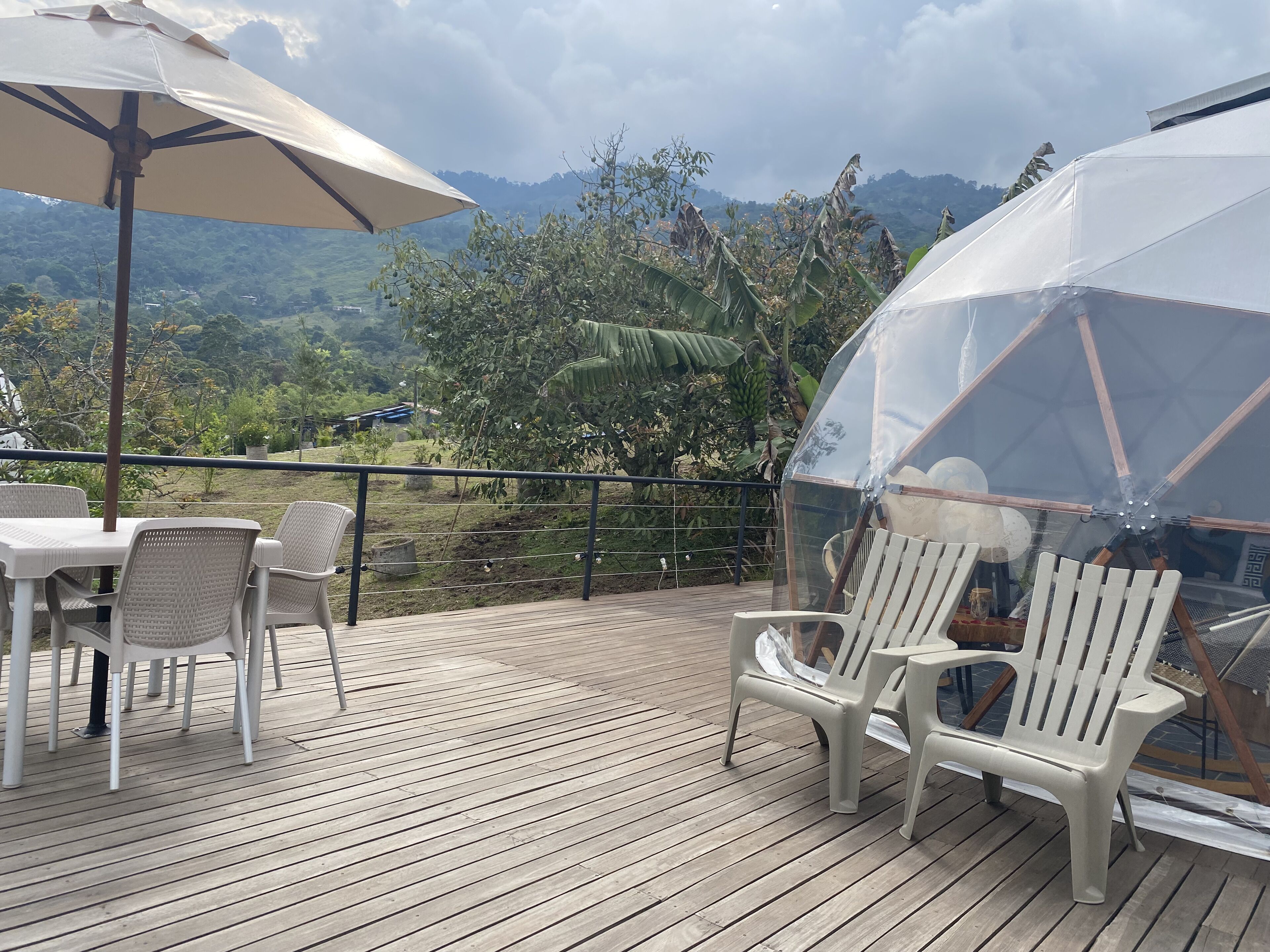 Una terraza de madera con sillas de plástico blanco y una mesa, un área cubierta con una cúpula transparente, y vistas a un paisaje montañoso.