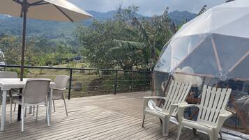 Una terraza de madera con sillas de plástico blanco y una mesa, un área cubierta con una cúpula transparente, y vistas a un paisaje montañoso.