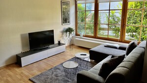 Un salon moderne doté d’un téléviseur à écran plat, d’un canapé gris et d’une grande fenêtre offrant une vue sur la verdure.