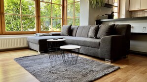 Un salon moderne avec un canapé gris, deux petites tables rondes et une grande fenêtre donnant sur un paysage verdoyant.