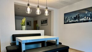 Un espace repas moderne avec une table blanche et des bancs noirs, un tableau représentant un paysage urbain accroché au mur et des lampes suspendues.