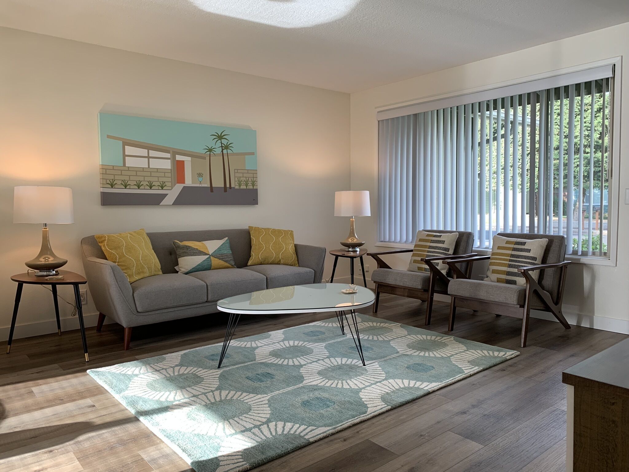 Una sala de estar moderna con un sofá gris, dos sillones, una mesa de centro y un tapete estampado. Hay un cuadro en la pared y una ventana con persianas.