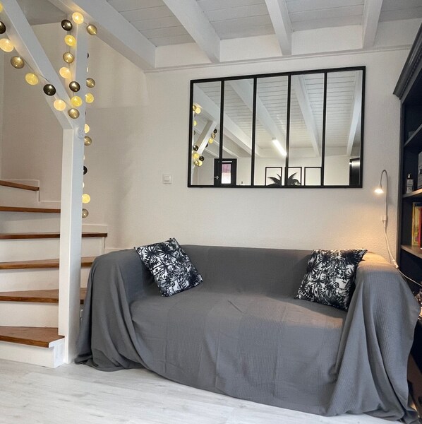 Un salon moderne avec un canapé gris, des coussins à motifs noir et blanc, un grand miroir et des guirlandes lumineuses suspendues au plafond.