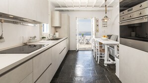 A modern kitchen with a central island, a dining area with a table and chairs, and a view through an open door to an outdoor patio. - Apanema Villas, Mykonos (Tourlos)