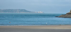 A beach with calm waters, a distant rocky coastline, and a small building on a hill.