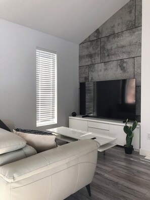 A modern living room with a sofa, a flat-screen TV mounted on a concrete wall, a potted plant, and a window with blinds. - Condo Orford (Orford)