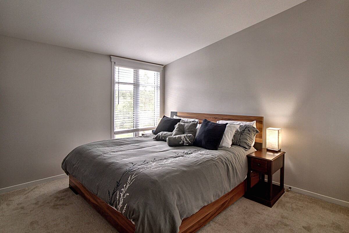 A bedroom with a large bed, a nightstand, a lamp, and a window with blinds.