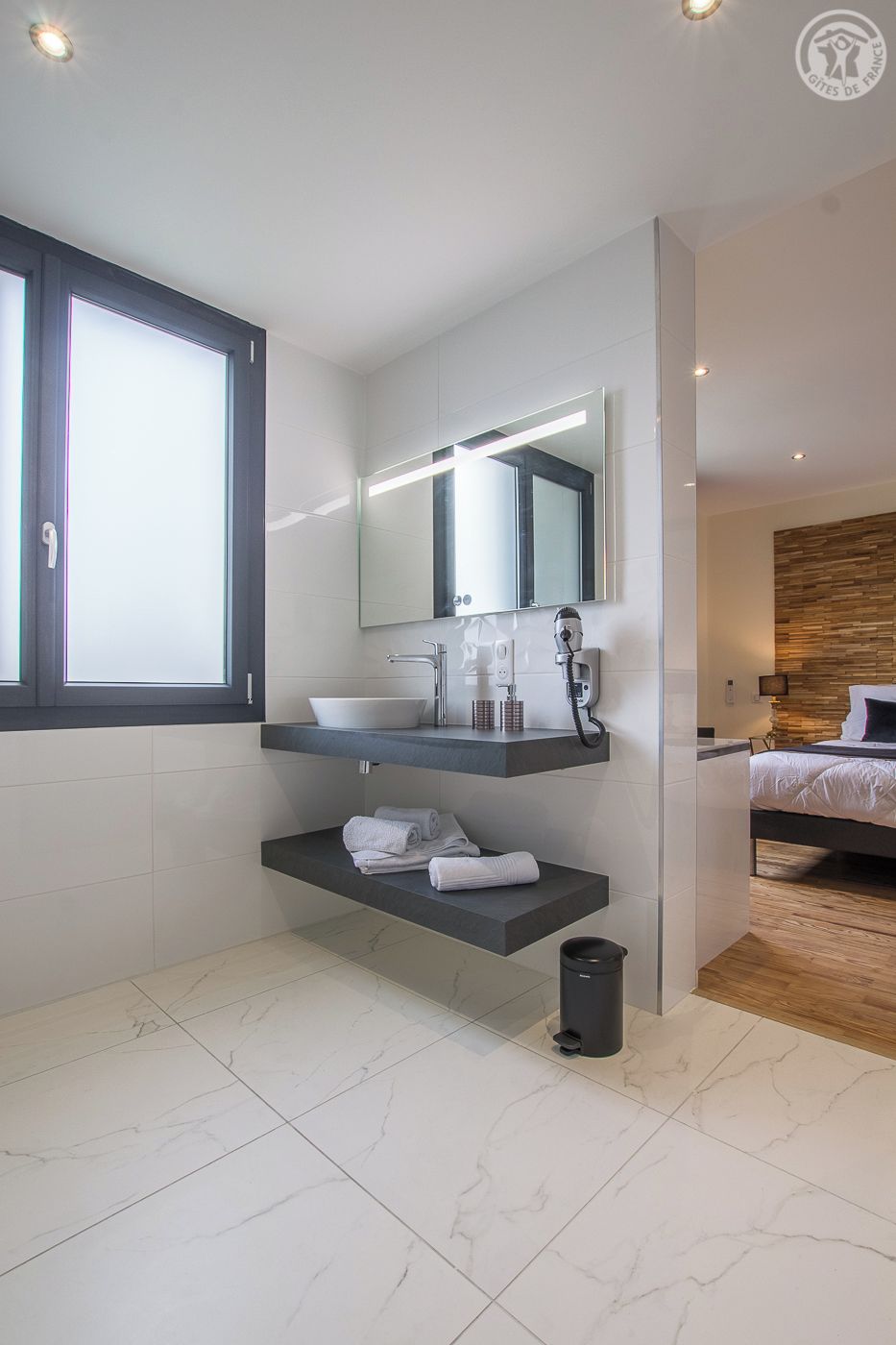 A modern bathroom with a large mirror, white marble tiles, a built-in sink, and a small trash bin.
