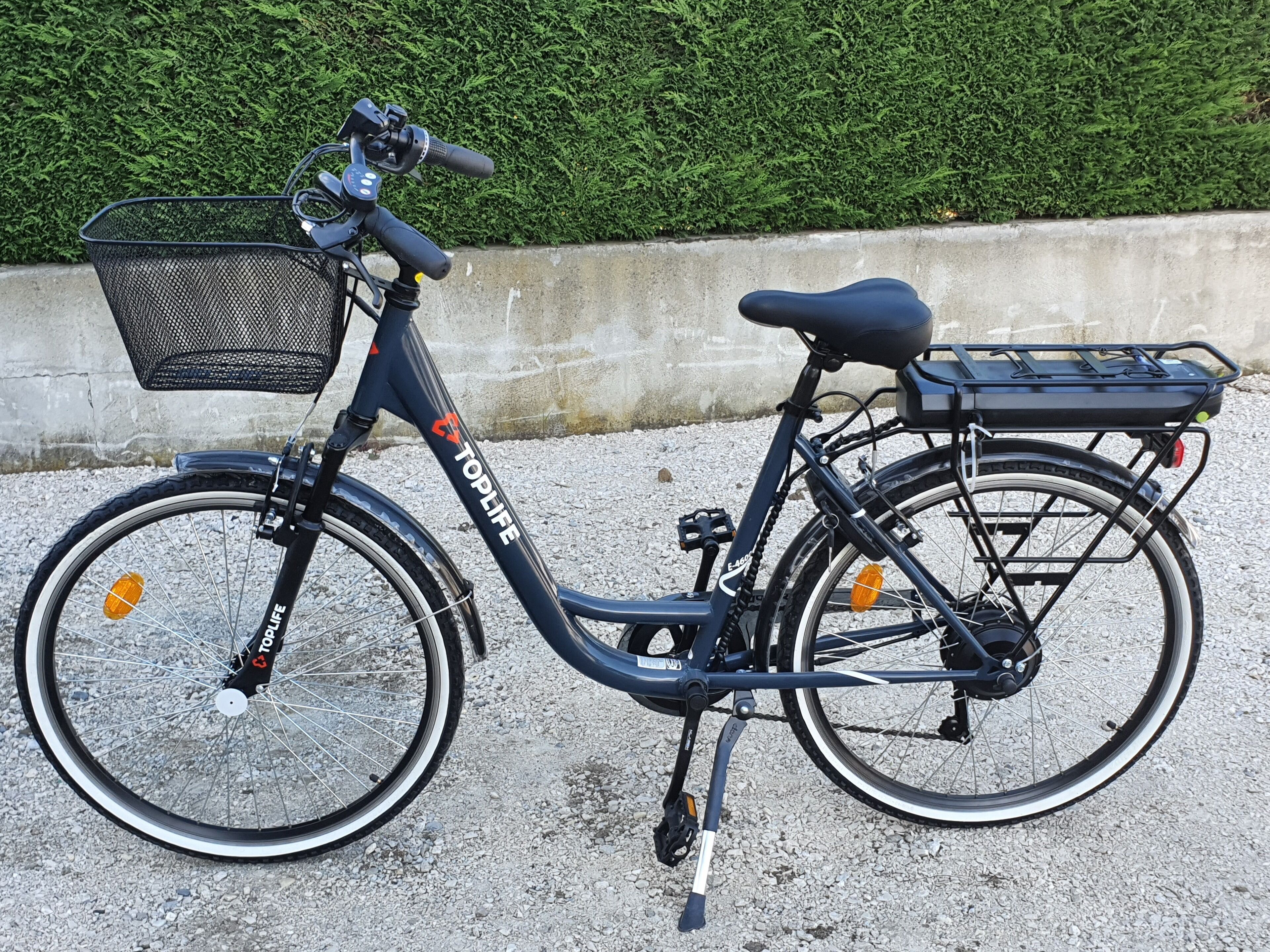 Una bicicleta eléctrica estacionada sobre grava, con canasta y portaequipajes trasero.