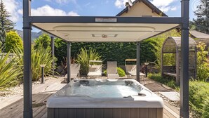 Ein Whirlpool unter einer überdachten Terrasse mit Liegestühlen, umgeben von Grünpflanzen und einem Haus im Hintergrund.