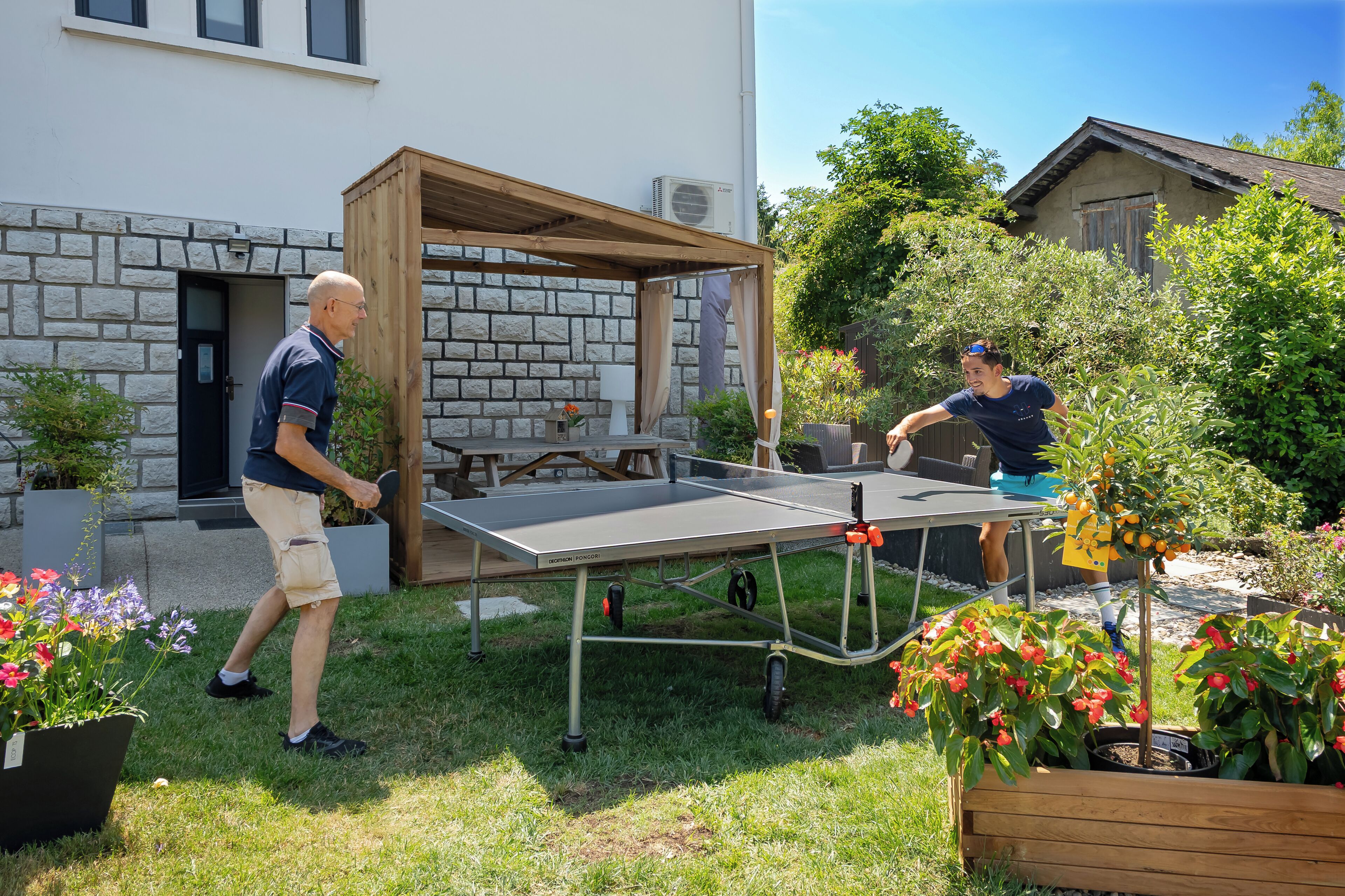 Mesa de ping pong montada en un jardín con un muro de piedra y una pérgola de madera.