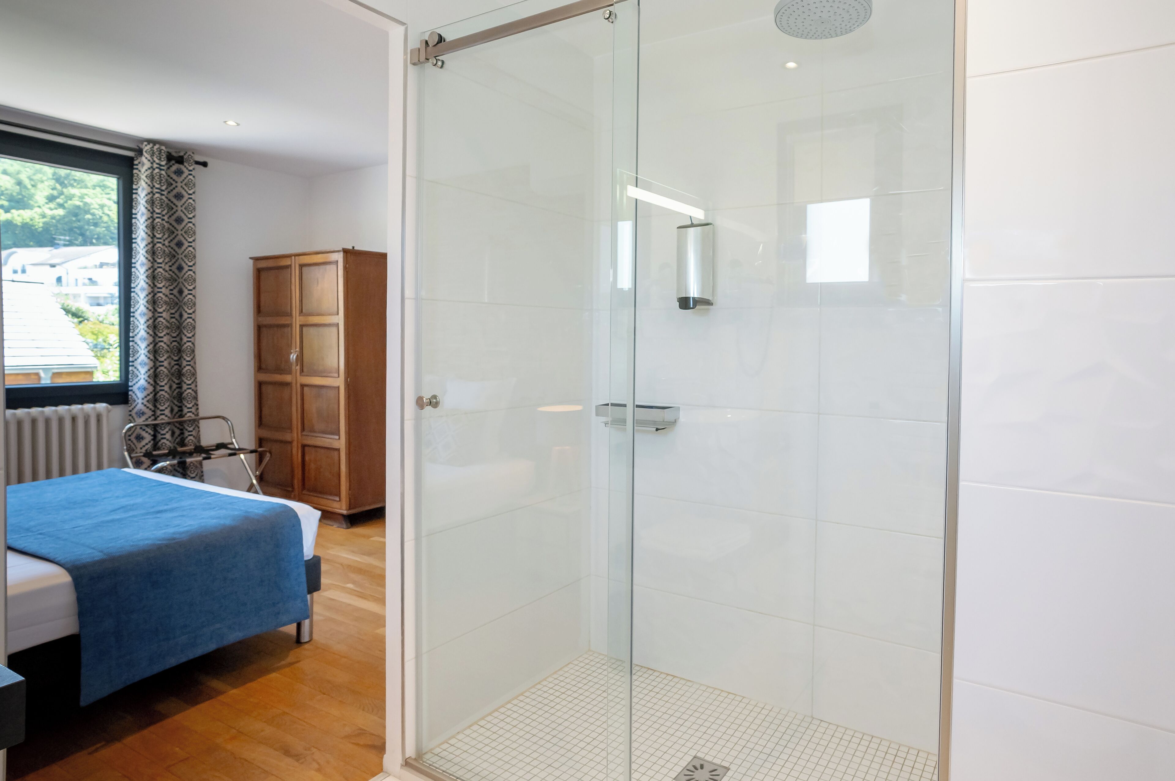 A modern bedroom with a glass shower enclosure, a bed with a blue blanket, and a wooden wardrobe.