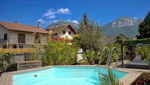 Ein Schwimmbecken mit Holzterrasse, umgeben von Pflanzen und Blick auf einen Berg.