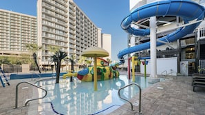 Uma área de piscina com escorregador, palmeiras e um edifício grande ao fundo.