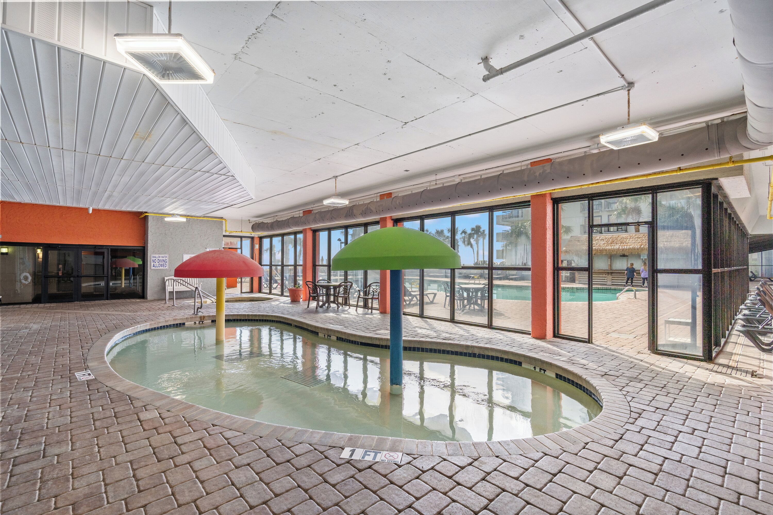 An indoor swimming pool with a brick floor, surrounded by glass walls and a poolside area with tables and chairs.